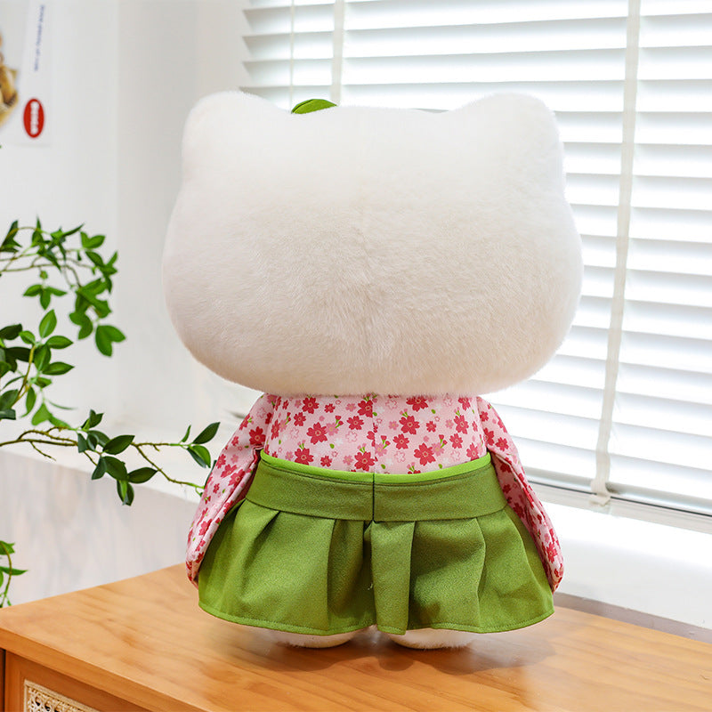 A plush toy with a large white furry head and body, wearing a pink floral top and green skirt, sitting on a wooden table in front of a window with white blinds.