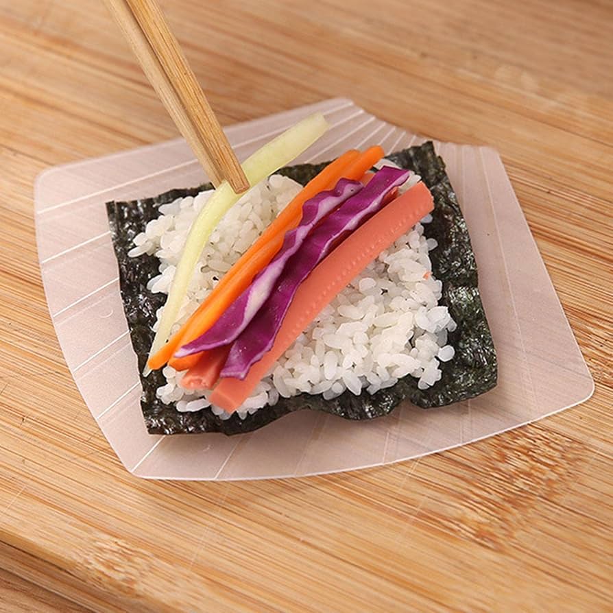 A square translucent plastic temaki sushi mold on a wooden surface, holding a sheet of seaweed with white rice and colorful vegetable strips including carrot, red cabbage, and cucumber, with chopsticks resting on top.