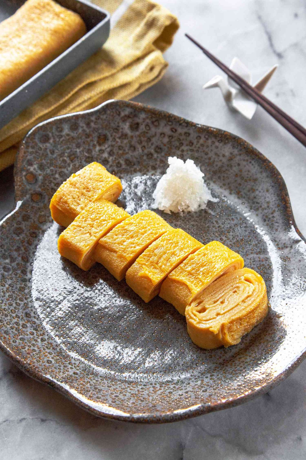 Sliced golden-yellow Japanese tempura or egg roll pieces served on a speckled gray ceramic plate with a small mound of white rice, set on a light marble surface with chopsticks and a baking pan in the background.
