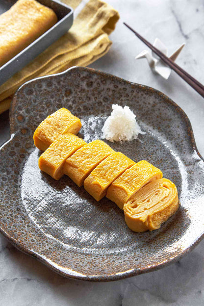 Sliced golden-yellow Japanese tempura or egg roll pieces served on a speckled gray ceramic plate with a small mound of white rice, set on a light marble surface with chopsticks and a baking pan in the background.